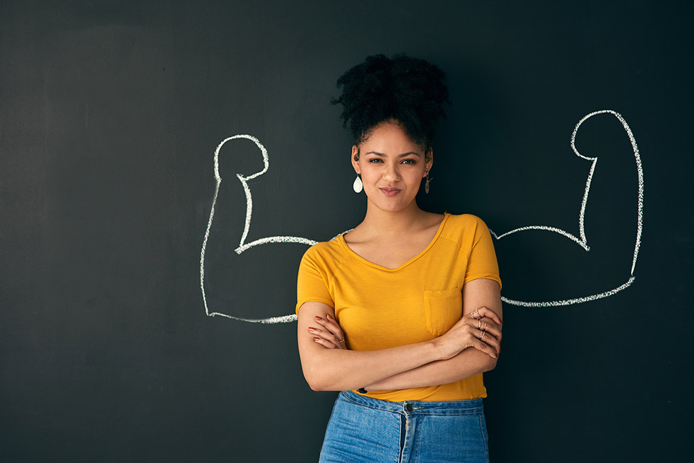 (image of a woman standing in front of a chalkboard with strong arms drawn on it behind her as if she's flexing her muscles)